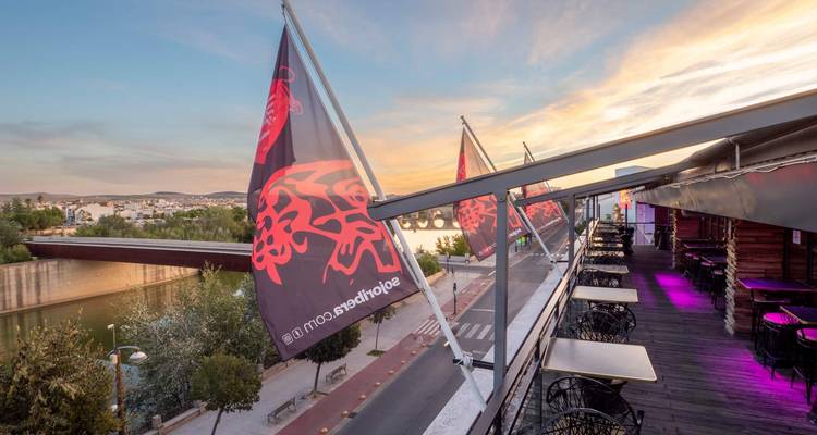 Vue du coucher de soleil depuis une terrasse sur le toit bordée de drapeaux et de tables de café surplombant une rivière et un pont.