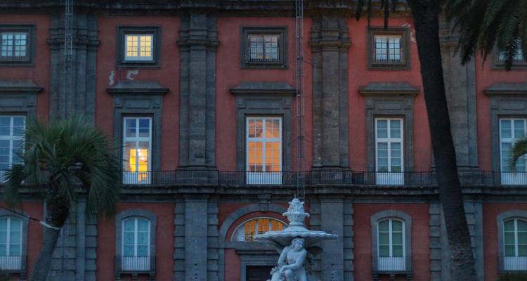 Historische rode zandsteen paleisgevel in het schemerlicht met verlichte ramen en een fonteinbeeldhouw ervoor.