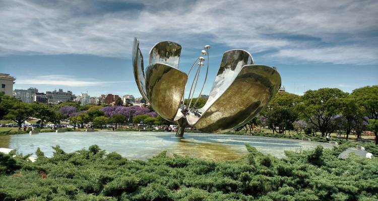 Weergave vanaf grondniveau van Floralis Genérica open met helder waterbassein en paarse jacarandabomen in bloei