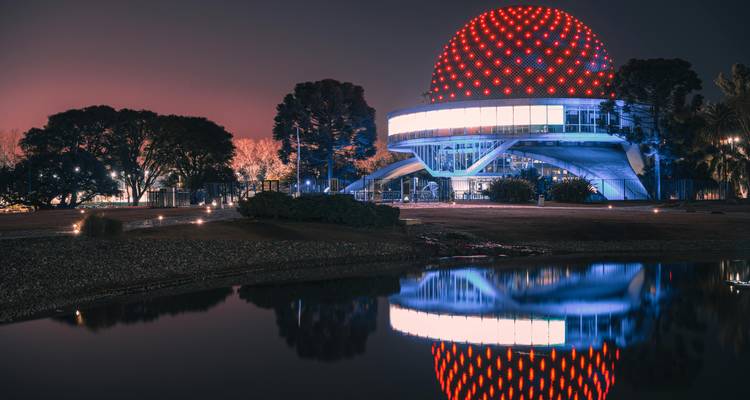 Nachtfoto van het verlichte Galileo Galilei Planetarium met zijn karakteristieke rode koepel gespiegeld in het stille water