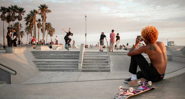 Scène dynamique au skatepark de Venice Beach avec des skateurs effectuant des figures acrobatiques et des spectateurs qui regardent au crépuscule.