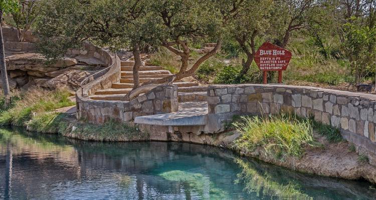 Piscine naturelle d'eau claire et bleue au printemps, entourée de murs en pierre, avec un panneau indiquant « Blue Hole », des arbres et des marches descendantes