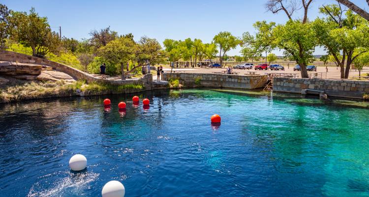 Piscine naturelle d'un bleu éclatant parsemée de bouées rouges et blanches, avec des personnes debout au bord en pierre sous un ciel ensoleillé
