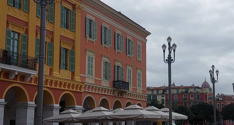 Bâtiment historique coloré avec des arches et des volets dans une place de ville méditerranéenne par un jour nuageux.