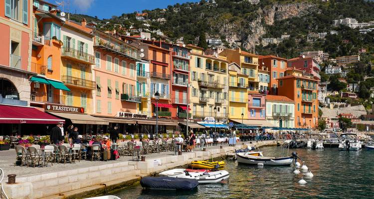 Bord de mer vibrant d'un village méditerranéen avec des maisons aux couleurs pastel, des terrasses de café, des bateaux et des collines en arrière-plan.