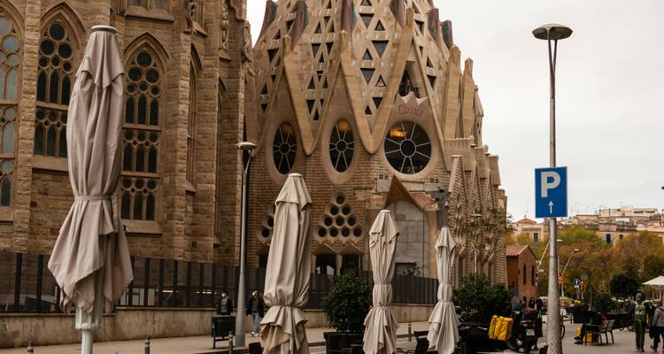 Außenansicht der kunstvollen Fassade der Sagrada Familia mit eingeklappten Regenschirmen und Fußgängern an einem bewölkten Tag