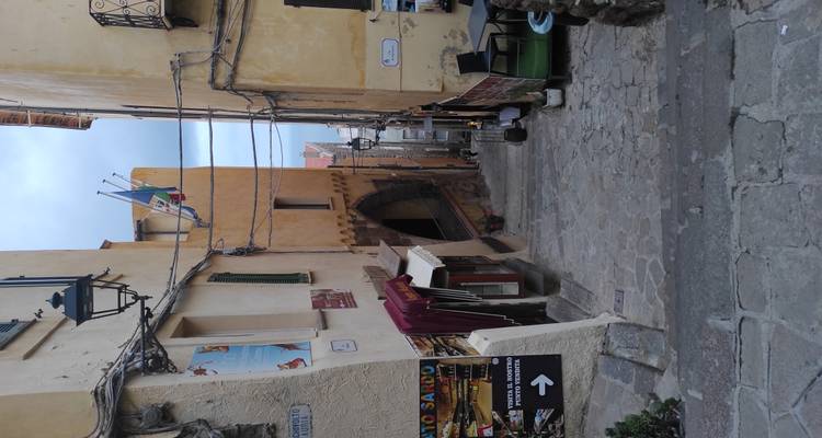 Ruelle en pierre étroite et escarpée dans une ville historique colorée de Sardaigne, bordée de drapeaux, de lanternes et de petits cafés, descendant vers la mer lointaine.