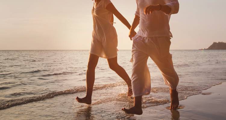 Couple en silhouette vêtu de blanc marchant pieds nus le long du rivage au coucher du soleil.