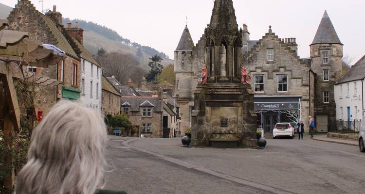 Femme aux cheveux gris observant une charmante place de village écossais historique avec un monument médiéval.