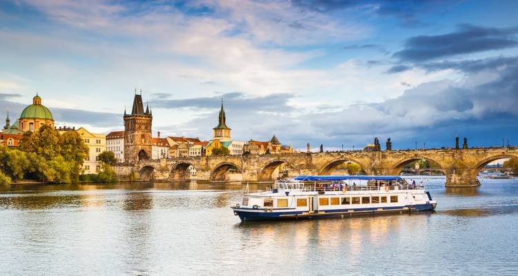Bote de tour pasando bajo el histórico Puente de Carlos con el horizonte de Praga al atardecer.