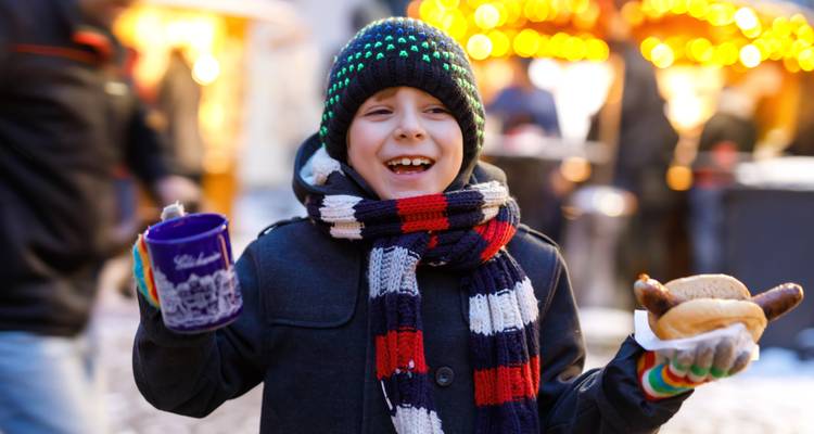 Lachendes Kind, das sich auf einem Weihnachtsmarkt warm eingepackt hat und ein heißes Getränk sowie eine Bratwurst hält.
