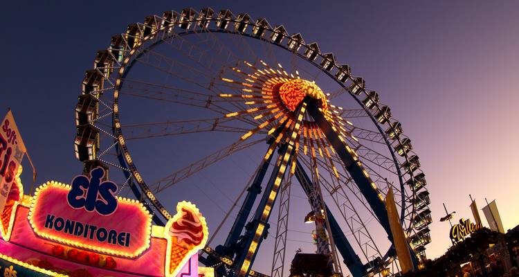 Das beleuchtete Riesenrad und Neon-Essbudenstände schaffen eine lebendige Jahrmarktszene in der Abenddämmerung.