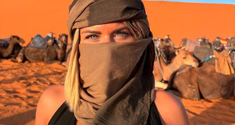 Vrouw met blauwe ogen en hoofddoek staat voor rustende kamelen op oranje woestijnduinen.