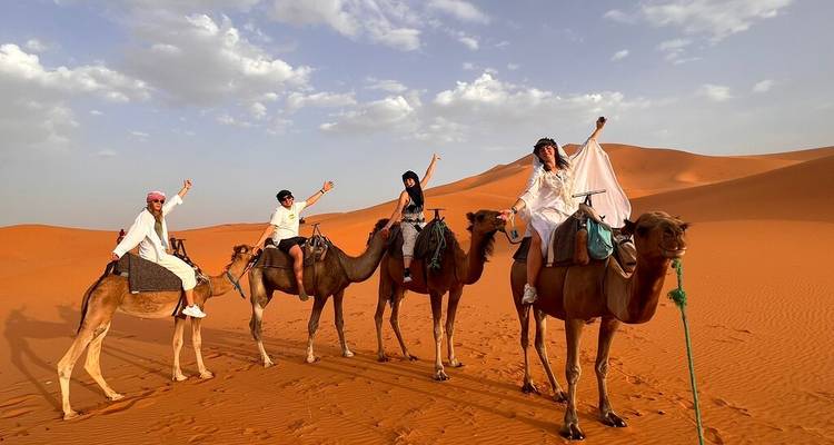 Reizigers juichen bovenop een kamelenkaravaan die over golvende Sahara-zandduinen rijdt onder verspreide wolken.