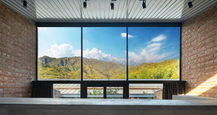 Chambre minimaliste moderne avec une fenêtre panoramique encadrant les montagnes luxuriantes et le ciel bleu.