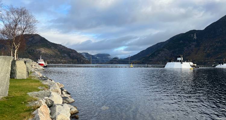 Port tranquille dans un fjord avec pont, bateaux et montagnes forestières environnantes sous un ciel nuageux brillant.