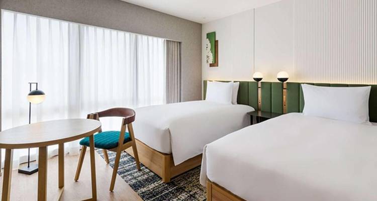 Habitación de hotel moderna y luminosa con dos camas individuales, muebles de madera clara y una gran ventana con cortinas.
