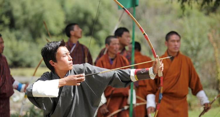 Bhutanische Männer in traditioneller Kleidung üben sich im Freien im Bogenschießen, ein Bogenschütze im Vordergrund spannt einen Bogen mit großer Konzentration.