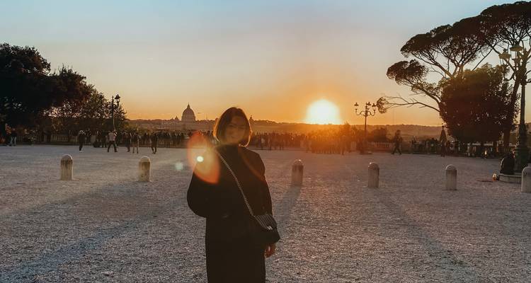 Silhouet van een vrouw die in een groot plein staat bij zonsondergang met de koepel van Sint-Pieter die aan de horizon gloeit.
