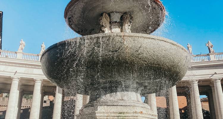 Close-up van een stenen fontein die water spuit op het Sint-Pietersplein met zuizenkolonnades op de achtergrond.