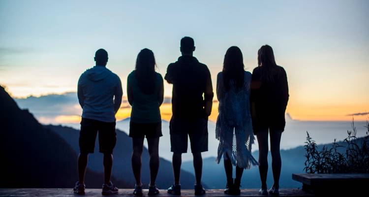 Des amis en silhouette se tiennent côte à côte, surplombant des vallées sombres et un ciel à l'aube aux tons pastel.
