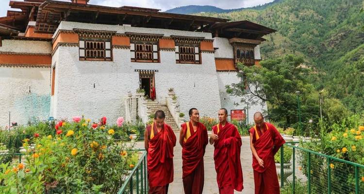 Four monks in red robes chat while walking in front of a white-walled Bhutanese dzong surrounded by gardens.