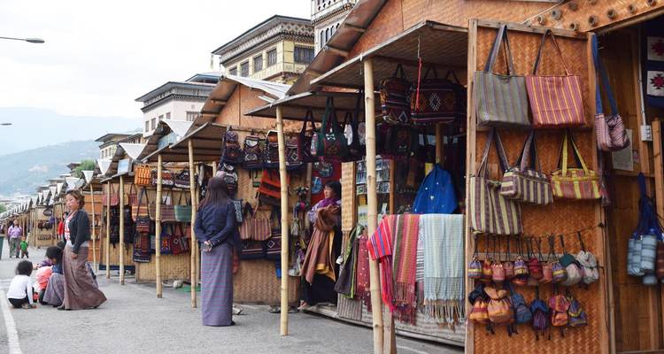 Row of outdoor market stalls selling colorful woven bags and fabrics on a Bhutanese street.
