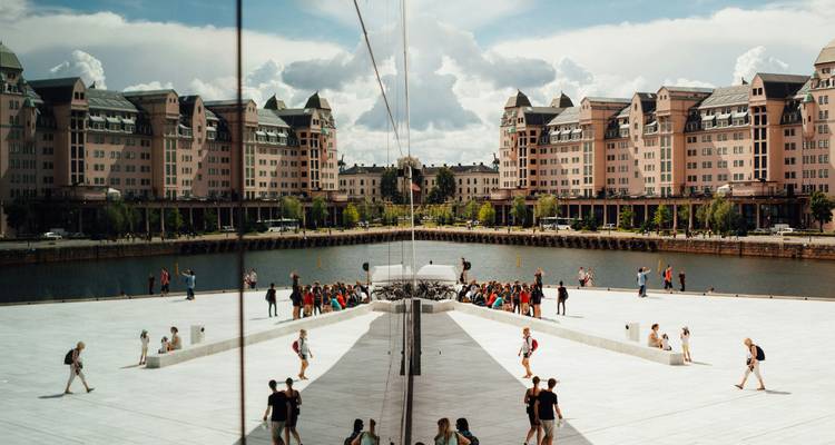 Des visiteurs sur le toit de l'Opéra d'Oslo avec une façade en miroir reflétant les deux hôtels roses de l'autre côté de l'eau.