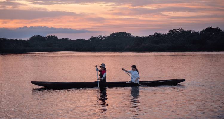Deux voyageurs pagaient un canoë étroit sur un lac calme face à un ciel de coucher de soleil coloré.