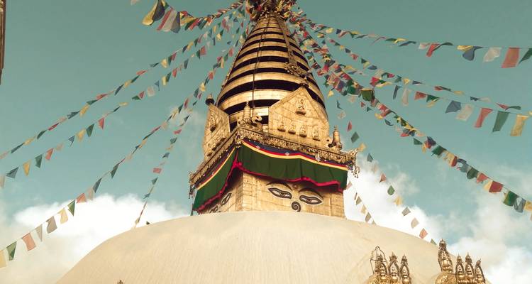 Aufwärtsblick auf die vergoldete Stupa-Spitze von Swayambhunath, umrahmt von Schnüren mehrfarbiger Gebetsfahnen und sanften Wolken.