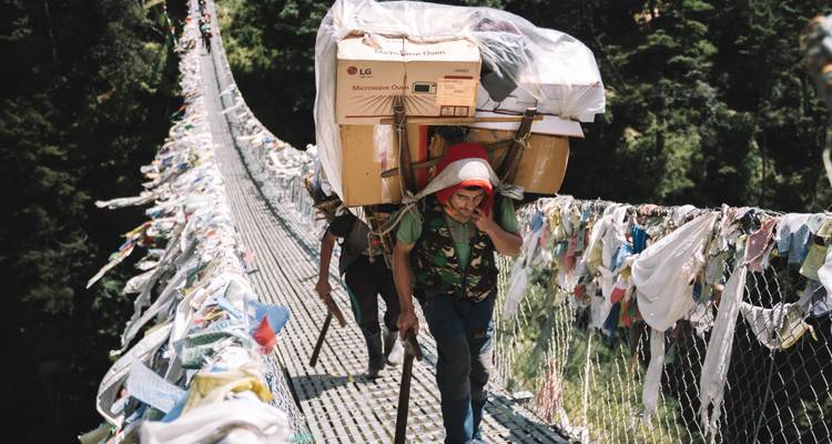 Träger, die mit großen Kisten beladen sind, durchqueren eine mit Gebetsfahnen geschmückte Hängebrücke in den Wäldern Nepals.