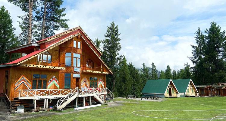 Un albergue alpino de madera y pequeñas cabañas en forma de A se encuentran en un campo de pasto bordeado por un bosque de pinos.