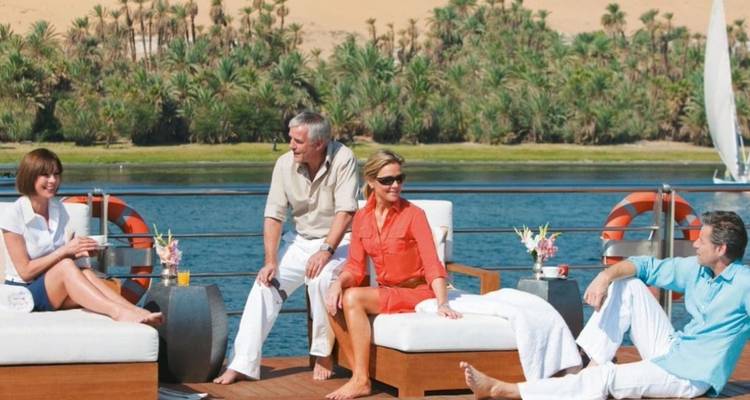 Des passagers détendus discutent sur des chaises longues à bord d'une croisière fluviale avec le paysage luxuriant du Nil en arrière-plan.
