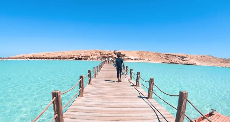 Un visiteur marche le long d'une longue jetée en bois surplombant une mer turquoise cristalline en direction d'une île sablonneuse.