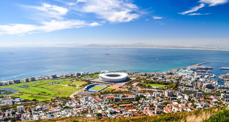 Vista aérea de Ciudad del Cabo con el Estadio de Ciudad del Cabo de forma circular, el paseo marítimo, la cuadrícula de la ciudad y la vasta bahía azul bajo cielos despejados.