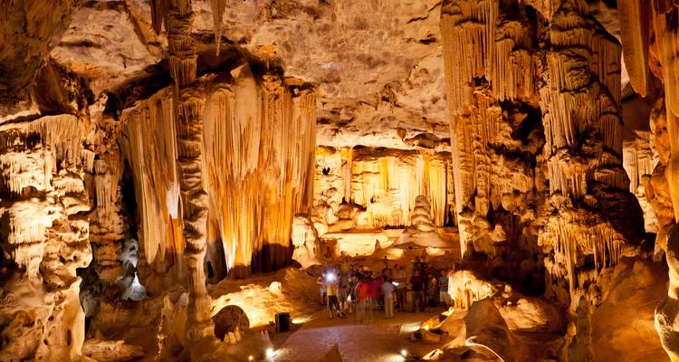 Cámara de cueva de piedra caliza iluminada con estalactitas y estalagmitas gigantes, visitantes explorando abajo con linternas.