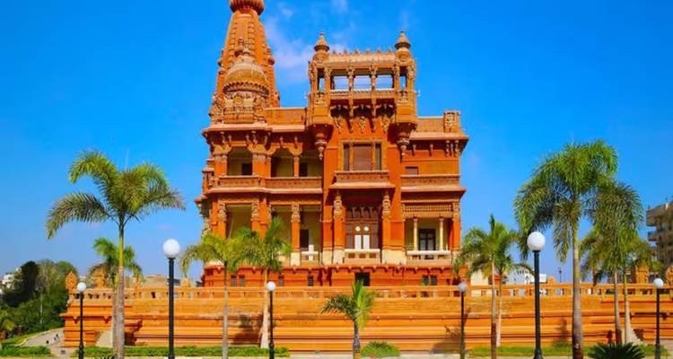 Palacio Baron Empain de color rojizo ornamentado enmarcado por palmeras contra un cielo despejado.