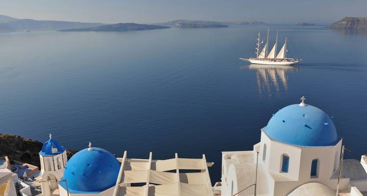 Blauwe koepels van Santorini met een zeilschip met hoge masten dat voorbijglijdt over het rustige calderawater