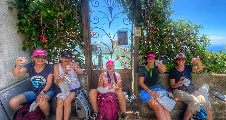 Cinq femmes vêtues de vêtements de randonnée portent un toast avec des collations et des boissons à côté d'une porte en fer forgé ouvragée donnant sur la mer.