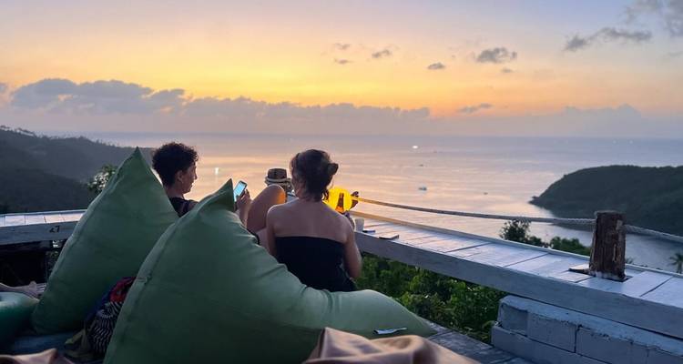 Los viajeros se relajan en puffs viendo una puesta de sol pastel sobre un vasto mar tropical y acantilados.