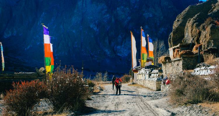 Deux trekkers marchent sur un sentier de terre bordé de drapeaux de prière colorés et de maisons en pierre dans l'Himalaya.