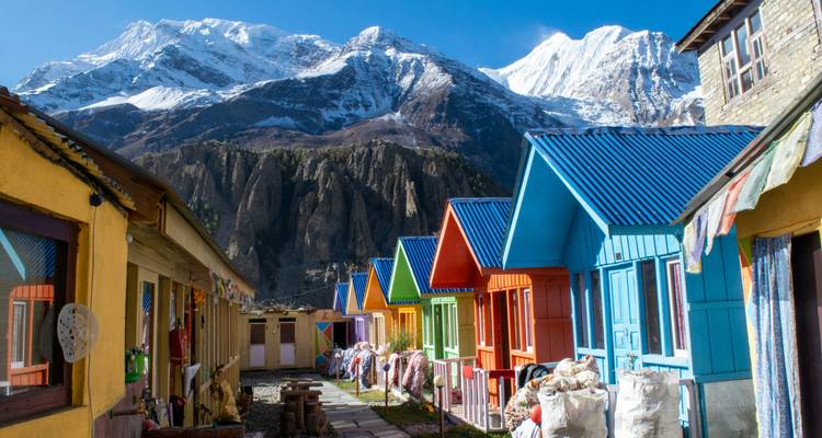 Une rangée de refuges de montagne peints de couleurs vives avec les pics dramatiques enneigés de l'Himalaya s'élevant derrière eux sous un ciel bleu dégagé.