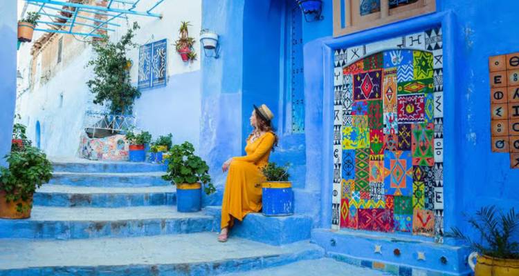 Une femme vêtue d'une robe jaune fluide se détend sur des marches peintes en bleu ornées de plantes en pot dans la médina de Chefchaouen.