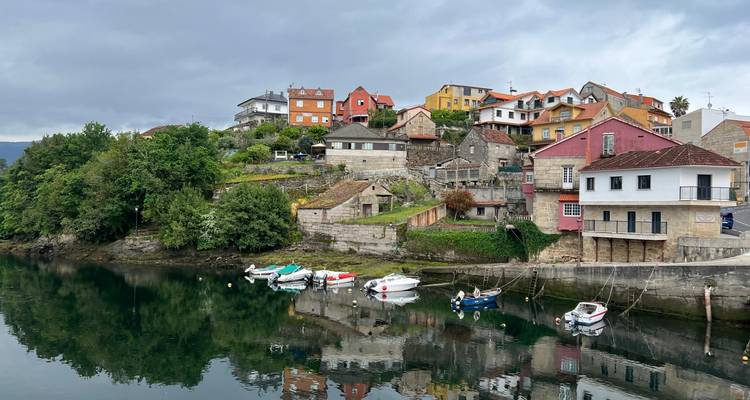 Village pittoresque en montagne avec des maisons colorées et de petits bateaux se reflétant sur une rivière calme sous un ciel gris.