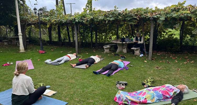 Les participants du cours de yoga sont allongés sur des tapis dans un jardin sous une pergola couverte de vignes.