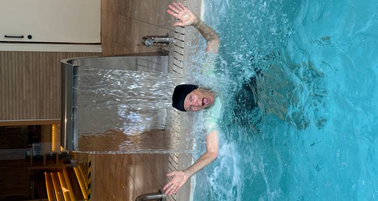 Un homme se tient joyeusement sous un jet de cascade dans une piscine intérieure, les bras levés.