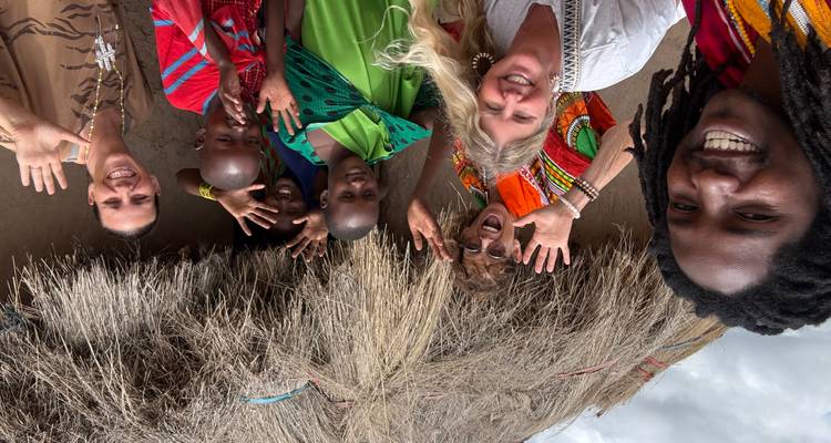 Reisende und Maasai-Gastgeber, die außerhalb einer Strohhütte für ein fröhliches Selfie lächeln.