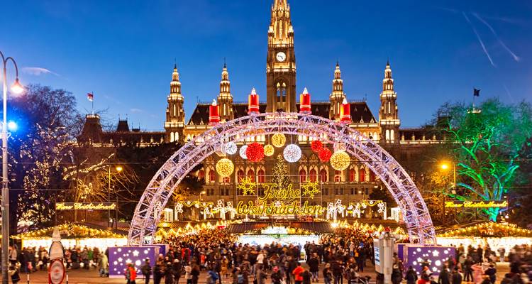 Belebter Weihnachtsmarkt am Wiener Rathausplatz mit funkelndem Bogen und beleuchteter Rathaus in der Abenddämmerung.