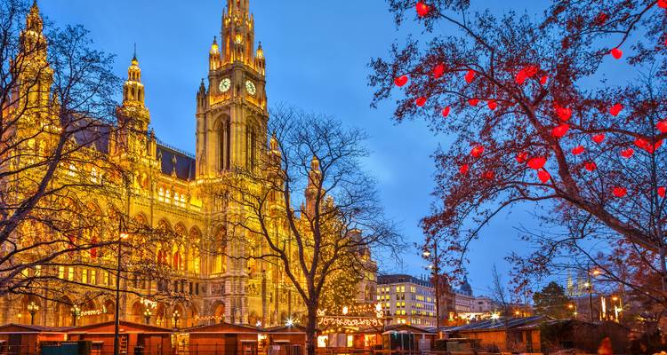 Das erleuchtete Wiener Rathaus hinter blattlosen Bäumen, die mit roten Herzornamenten geschmückt sind, am Abend.