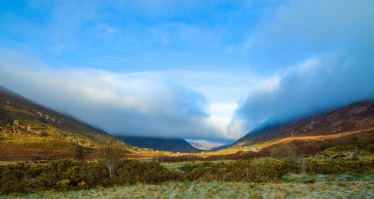 Valle de las Tierras Altas escocesas envuelto en niebla con laderas iluminadas por el sol bajo un cielo azul y nubes ondulantes.